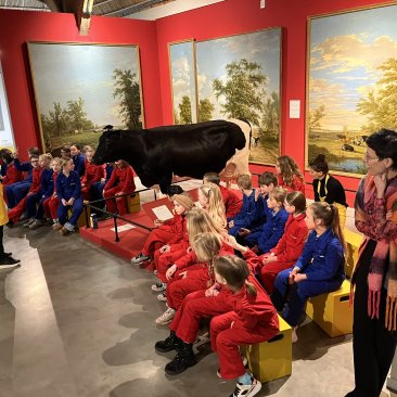 Kinderen in overall luisterend naar het introductieverhaal van een vrijwilligster voor de educatieles