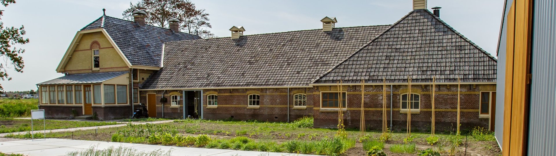 Foto van de voorkant van de W.C. de Groot boerderij waar het Fries Landbouwmuseum in gevestigd is. Op de voorgrond zijn opkomende gewassen te zien in de moestuin van het museum.