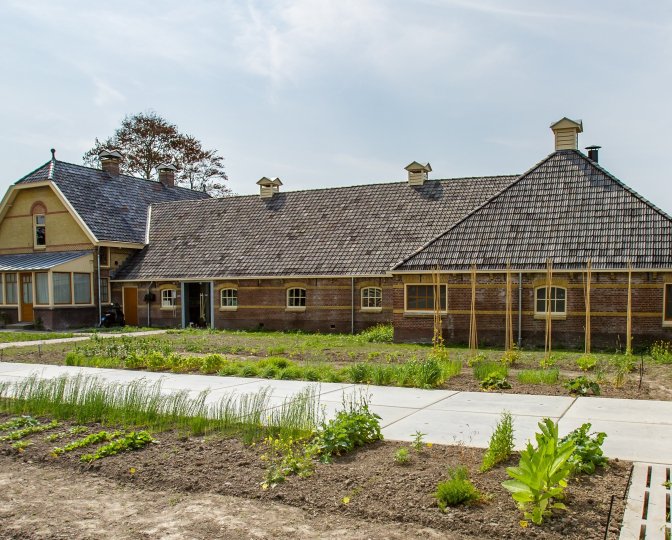 Foto van de voorkant van de W.C. de Groot boerderij waar het Fries Landbouwmuseum in gevestigd is. Op de voorgrond zijn opkomende gewassen te zien in de moestuin van het museum.