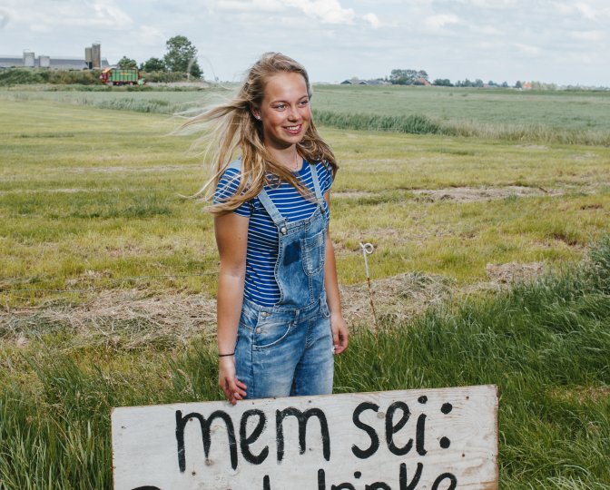 Een persoon in een weiland met een bord waarop staat 'mem sei: goed drinke mei dit waar!'